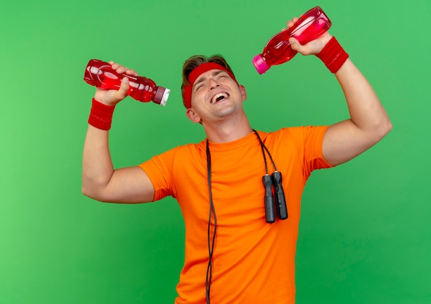 Freudiger junger hübscher sportlicher mann, der stirnband und armbänder mit springseil um den hals trägt, der wasserflaschen lokalisiert auf grün anhebt