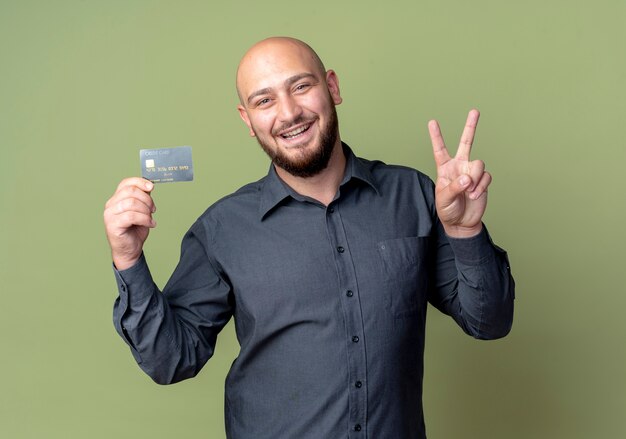 Freudiger junger kahlköpfiger Callcenter-Mann, der Kreditkarte hält Friedenszeichen lokalisiert auf olivgrün