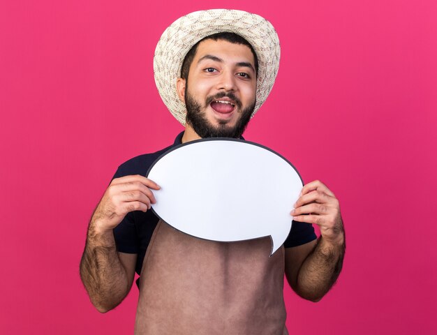 freudiger junger kaukasischer männlicher Gärtner, der Gartenhut hält, der Sprachballon hält, der auf rosa Wand mit Kopienraum lokalisiert wird