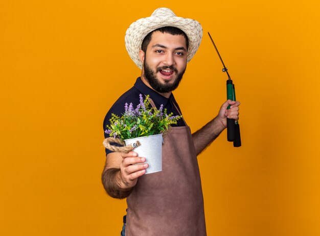 freudiger junger kaukasischer männlicher Gärtner, der Gartenhut trägt, steht seitlich und hält Gartenschere und Blumentopf lokalisiert auf orange Wand mit Kopienraum