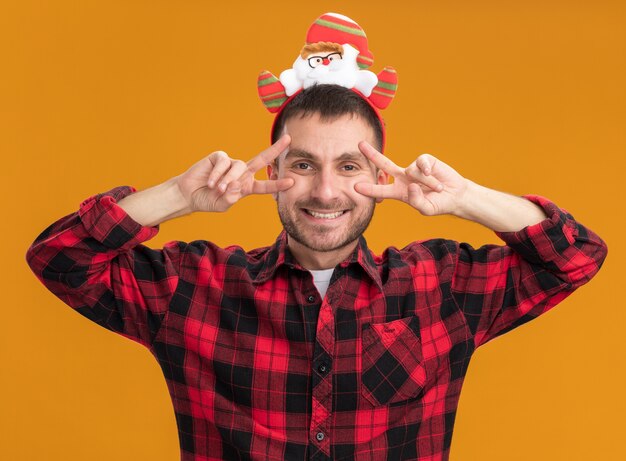 Freudiger junger kaukasischer Mann, der Weihnachtsmann-Stirnband trägt, das V-Zeichensymbole nahe den Augen lokalisiert auf orange Wand zeigt