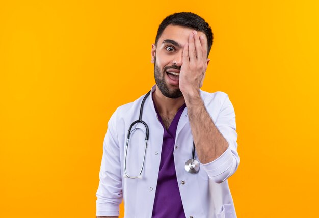 Freudiger junger männlicher Arzt, der medizinisches Kleid des Stethoskops trägt, bedeckte Augen mit Hand auf isolierter gelber Wand