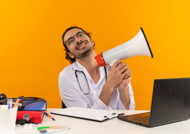 Freudiger junger männlicher Arzt mit medizinischer Brille, die medizinische Robe mit Stethoskop sitzt, sitzt am Schreibtischarbeit auf Laptop mit medizinischen Werkzeugen, die Lautsprecher auf lokalisiertem gelbem Hintergrund mit Kopienraum halten