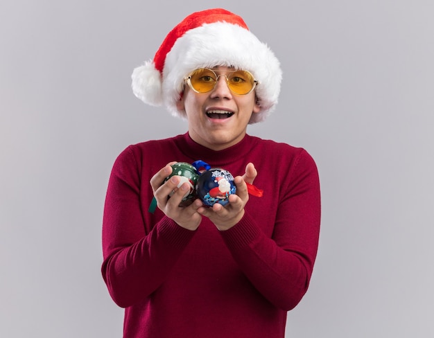 Freudiger junger Mann, der Weihnachtsmütze mit Brille trägt, die Weihnachtsbaumkugeln lokalisiert auf weißem Hintergrund hält