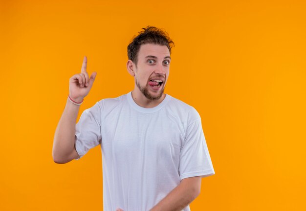 Freudiger junger Mann, der weißes T-Shirt trägt, das Zunge zeigt, zeigt oben auf isolierter orange Wand