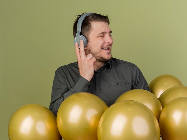 Freudiger junger Party-Typ, der schwarzes Hemd und Kopfhörer trägt, die unter den auf olivgrün isolierten Luftballons stehen