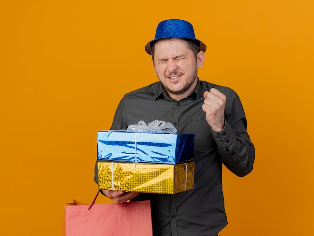 Freudiger junger Party-Typ mit geschlossenen Augen, der blauen Hut trägt, der Geschenkboxen und Tasche hält, die ja Geste lokalisiert auf Orange zeigt