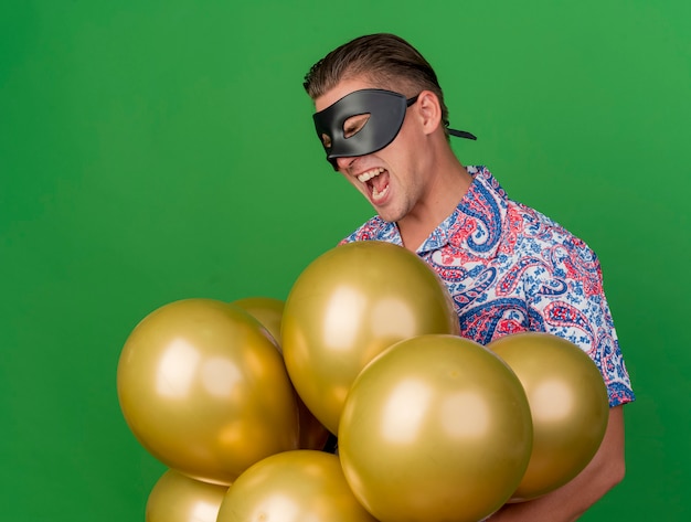 Freudiger junger Party-Typ mit geschlossenen Augen, der Maskerade-Augenmaske hält, die Luftballons lokalisiert auf Grün hält