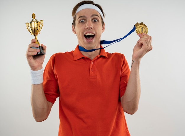 Freudiger junger sportlicher Kerl, der Stirnband mit Armband hält Gewinnerpokal mit Medaille lokalisiert auf weißer Wand trägt
