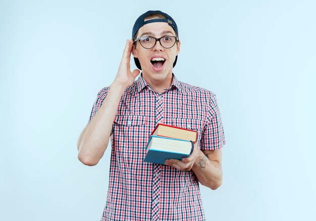 Freudiger junger Studentjunge, der Brille und Kappe trägt Bücher hält und Hand auf Ohr legt