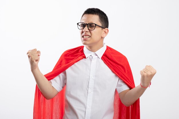 Freudiger junger Superheldenjunge im roten Umhang, der Brille tut, die ja Geste tut, die Seite lokalisiert auf weißem Hintergrund betrachtet