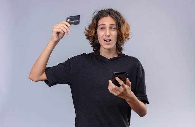 Freudiger Kerl mit langen Haaren im schwarzen T-Shirt, der ein Telefon und Bankard auf weißer Wand hält