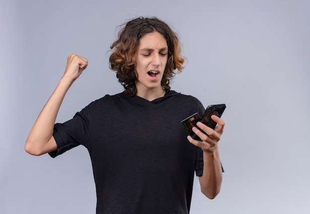 Freudiger kerl mit langen haaren im schwarzen t-shirt, der ein telefon und eine karte auf weißer wand hält