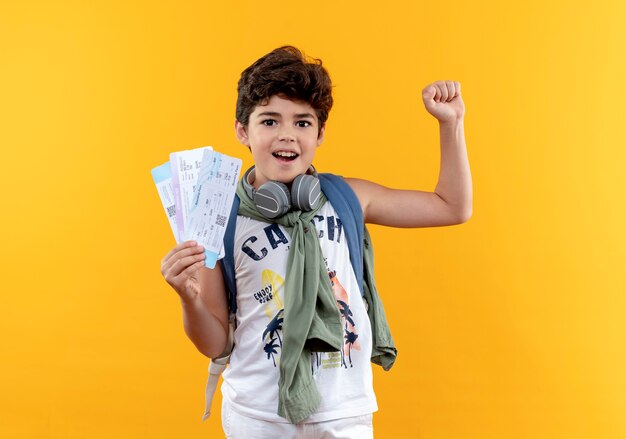 Freudiger kleiner Schuljunge, der Rückentasche und Kopfhörer trägt, die Karten halten und Ja-Geste lokalisiert auf gelbem Hintergrund zeigen