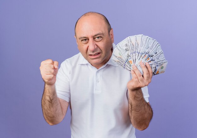 Freudiger lässiger reifer Geschäftsmann, der Geld und geballte Faust hält, die ja Geste lokalisiert auf lila Hintergrund tut