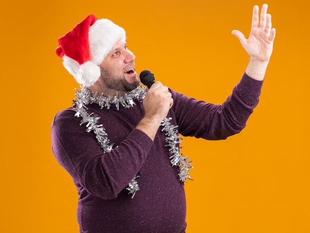 Kostenloses Foto freudiger mann mittleren alters, der weihnachtsmütze und lametta-girlande um den hals hält, der mikrofon hält, das oben hängendes handgesang lokalisiert auf orangefarbenem hintergrund schaut