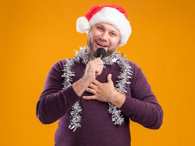 Freudiger Mann mittleren Alters, der Weihnachtsmütze und Lametta-Girlande um den Hals trägt, spricht in Mikrofon, das Kamera betrachtet, die Hand auf Brust lokalisiert auf orange Hintergrund hält