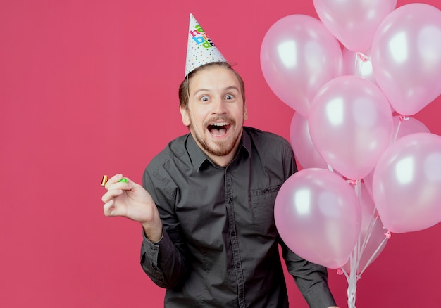 Freudiger schöner Mann in der Geburtstagskappe steht mit Heliumballons, die Pfeife lokalisiert auf rosa Wand halten