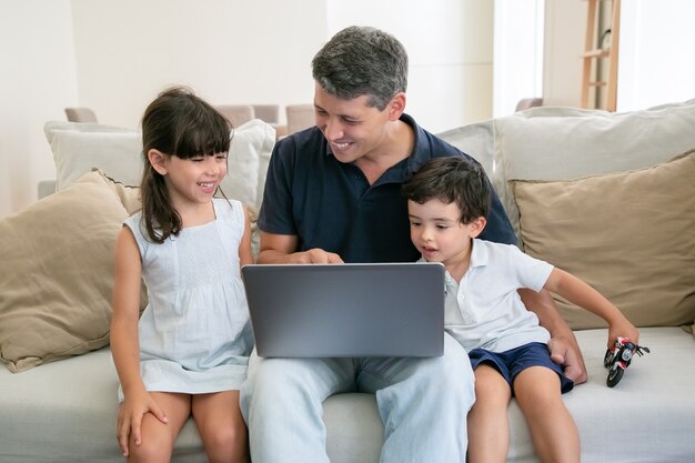 Freudiger Vater, der auf Bildschirm zeigt und glücklichen Kindern Inhalt auf Laptop zeigt.