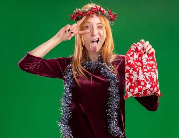 Kostenloses Foto freudiges blinzelndes junges schönes mädchen, das rotes kleid mit kranz und girlande am hals hält, die weihnachtstasche hält zunge zeigt und friedensgeste lokalisiert auf grüner wand tut