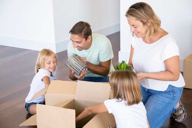 Freudiges Familienpaar und entzückende Mädchen, die in eine neue Wohnung ziehen, Spaß haben, während sie Dinge in einer neuen Wohnung auspacken, auf dem Boden sitzen und Gegenstände aus offenen Kisten nehmen