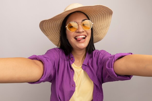 Freudiges hübsches brünettes kaukasisches Mädchen in der Sonnenbrille mit Strandhut steckt Zunge heraus und gibt vor, Kamera zu halten, die selfie auf Weiß nimmt