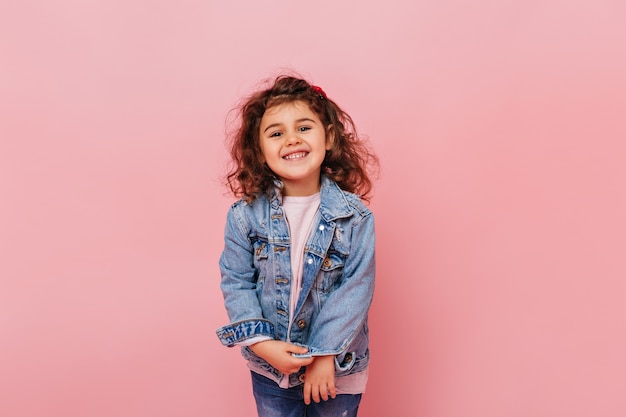 Freudiges jugendliches Kind mit dem lockigen Haar, das an Kamera lacht. Studioaufnahme des sorglosen kleinen Mädchens lokalisiert auf rosa Hintergrund.