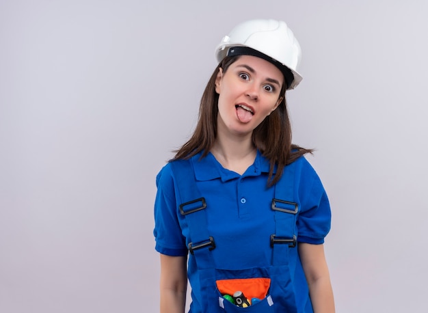Freudiges junges Baumeistermädchen mit weißem Sicherheitshelm und blauer Uniform betrachtet Kamera auf lokalisiertem weißem Hintergrund mit Kopienraum