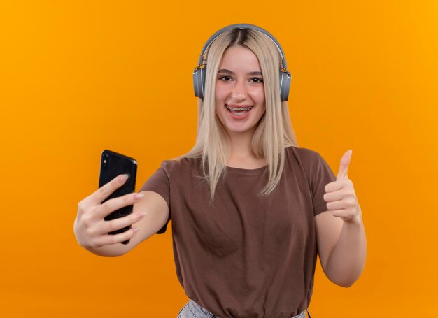 Freudiges junges blondes Mädchen, das Kopfhörer in Zahnspangen trägt, die Handy halten und Daumen oben auf lokalisiertem orangefarbenem Raum zeigen