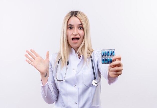 Freudiges junges blondes Mädchen des Arztes, das Stethoskop und medizinisches Kleid in Zahnspange trägt, die Pillen zur Kamera auf isolierter weißer Wand heraushalten