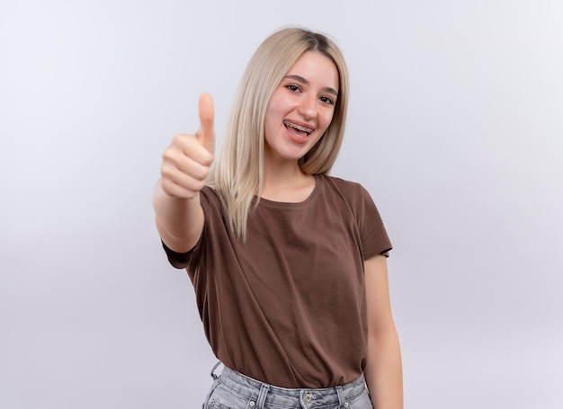 Freudiges junges blondes Mädchen in Zahnspangen, die Daumen oben auf lokalisiertem Leerraum mit Kopienraum zeigen