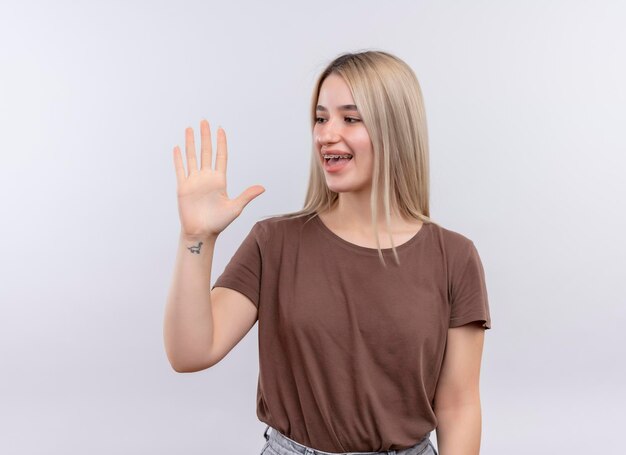 Freudiges junges blondes Mädchen in Zahnspangen, die fünf mit der Hand zeigen und es auf isoliertem weißem Raum mit Kopienraum betrachten