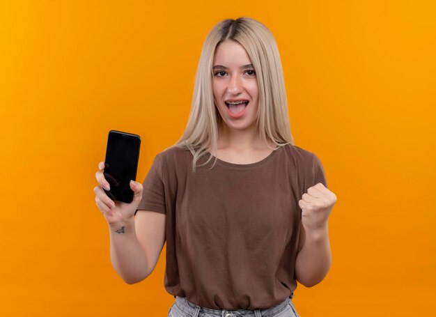 Freudiges junges blondes Mädchen in Zahnspangen, die Handy mit erhabener Faust auf lokalisiertem orange Raum mit Kopienraum halten