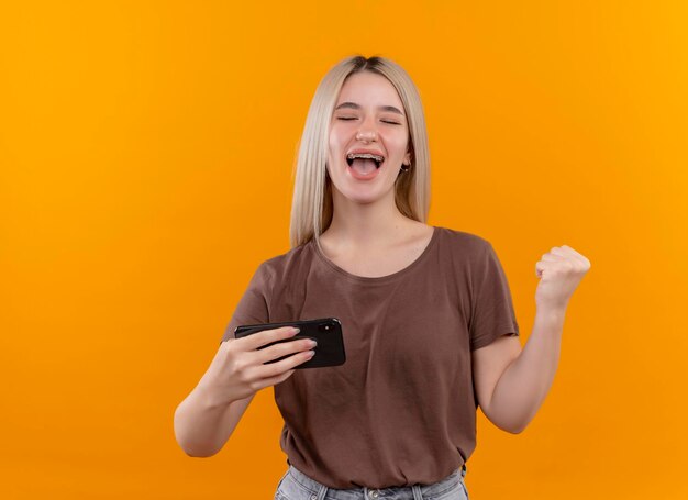 Freudiges junges blondes Mädchen in Zahnspangen, die Handy mit erhabener Faust und geschlossenen Augen auf lokalisiertem orange Raum mit Kopienraum halten