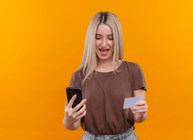 Freudiges junges blondes Mädchen in Zahnspangen, die Handy und Kreditkarte auf lokalisiertem orange Raum mit Kopienraum halten