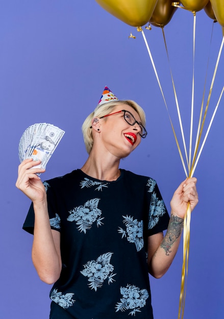 Freudiges junges blondes Partygirl, das Brille und Geburtstagskappe hält, die Ballons und Geld hält, die Ballons lokalisiert auf lila Hintergrund betrachten