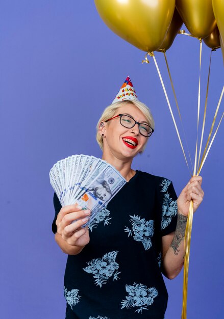 Freudiges junges blondes Partygirl, das Brille und Geburtstagskappe hält, die Ballons und Geld hält, die Seite lokalisiert auf lila Hintergrund betrachten
