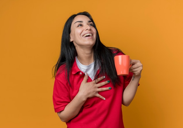 Freudiges junges brünettes kaukasisches Mädchen, das rotes Hemd trägt, legt Hand auf Brust und hält Tasse, die Seite betrachtet, die auf orange Wand lokalisiert ist