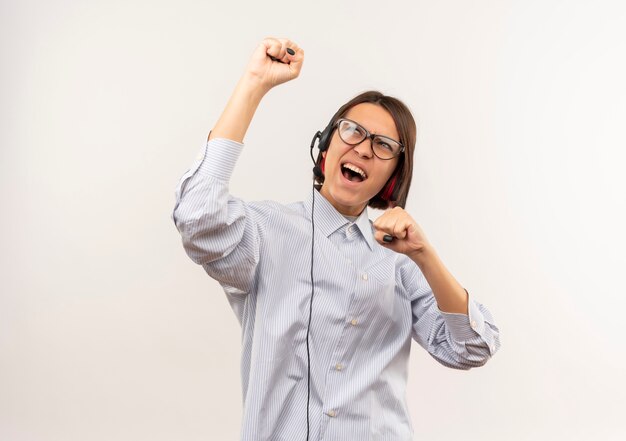 Freudiges junges Callcenter-Mädchen, das eine Brille und ein Headset trägt, das die auf Weiß lokalisierte Faust anhebt und betrachtet