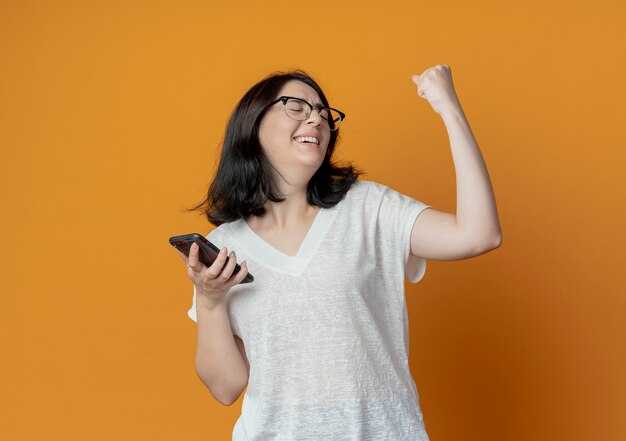 Freudiges junges hübsches kaukasisches Mädchen, das eine Brille trägt, die Handy hält und Faust gestikuliert, die ja mit geschlossenen Augen auf orange Hintergrund mit Kopienraum isoliert gestikuliert