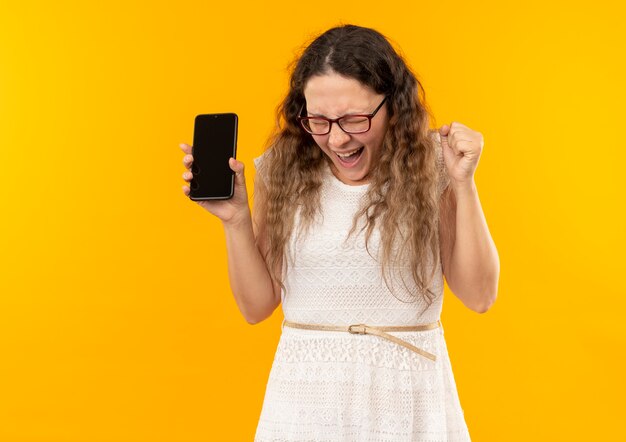 Freudiges junges hübsches Schulmädchen, das Brille und Rückentasche hält, die Handy-Faust mit geschlossenen Augen hält, die auf Gelb lokalisiert werden