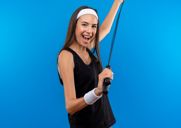 Freudiges junges hübsches sportliches Mädchen, das Stirnband und Armband hält Springseil lokalisiert auf blauem Raum trägt