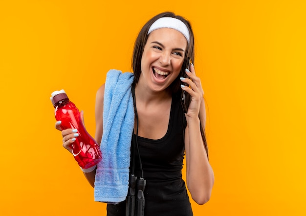 Freudiges junges hübsches sportliches Mädchen, das Stirnband und Armband hält Wasserflasche hält, die am Telefon spricht, das Seite betrachtet