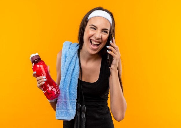 Freudiges junges hübsches sportliches Mädchen, das Stirnband und Armband hält Wasserflasche hält, die am Telefon spricht, das Seite betrachtet