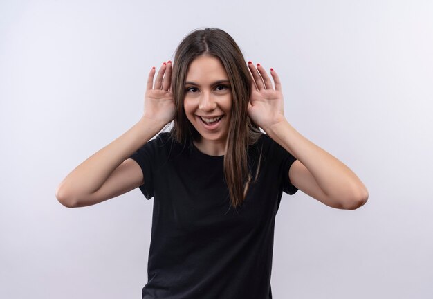 Freudiges junges kaukasisches Mädchen, das schwarzes T-Shirt trägt, legte Hände um Ohren auf isolierte weiße Wand
