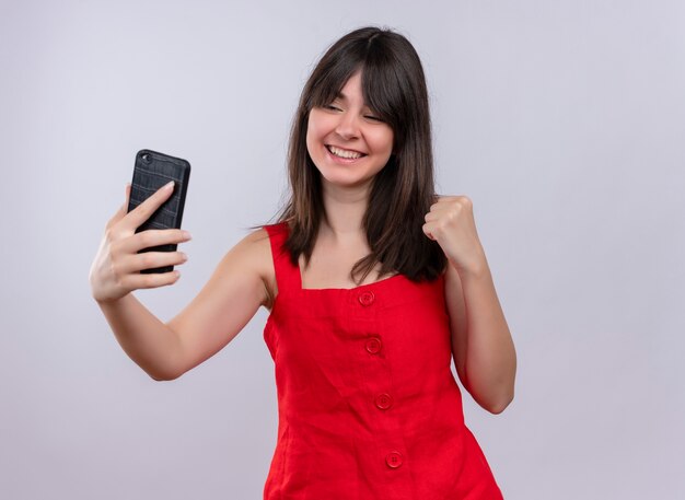 Freudiges junges kaukasisches Mädchen, das Telefon hält und Faust erhöht, die Telefon auf lokalisiertem weißem Hintergrund betrachtet