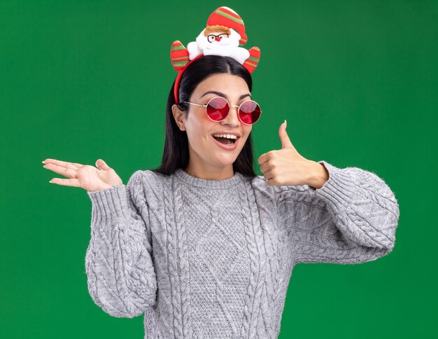 Freudiges junges kaukasisches Mädchen, das Weihnachtsmann-Stirnband mit Brille trägt, die Kamera zeigt, die leere Hand und Daumen oben auf grünem Hintergrund zeigt