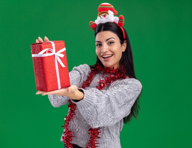 Freudiges junges kaukasisches Mädchen, das Weihnachtsmann-Stirnband und Lametta-Girlande um den Hals trägt, der Geschenkpaket in Richtung Kamera ausdehnt, die Kamera lokalisiert auf grünem Hintergrund betrachtet