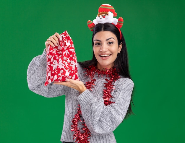 Freudiges junges kaukasisches Mädchen, das Weihnachtsmann-Stirnband und Lametta-Girlande um den Hals trägt, der Weihnachtsgeschenksack lokalisiert auf grüner Wand mit Kopienraum hält