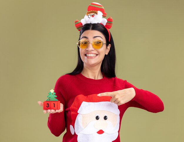 Freudiges junges kaukasisches Mädchen, das Weihnachtsmann-Stirnband und Pullover mit Gläsern hält, die auf Weihnachtsbaumspielzeug mit Datum lokalisiert auf olivgrüner Wand halten und zeigen
